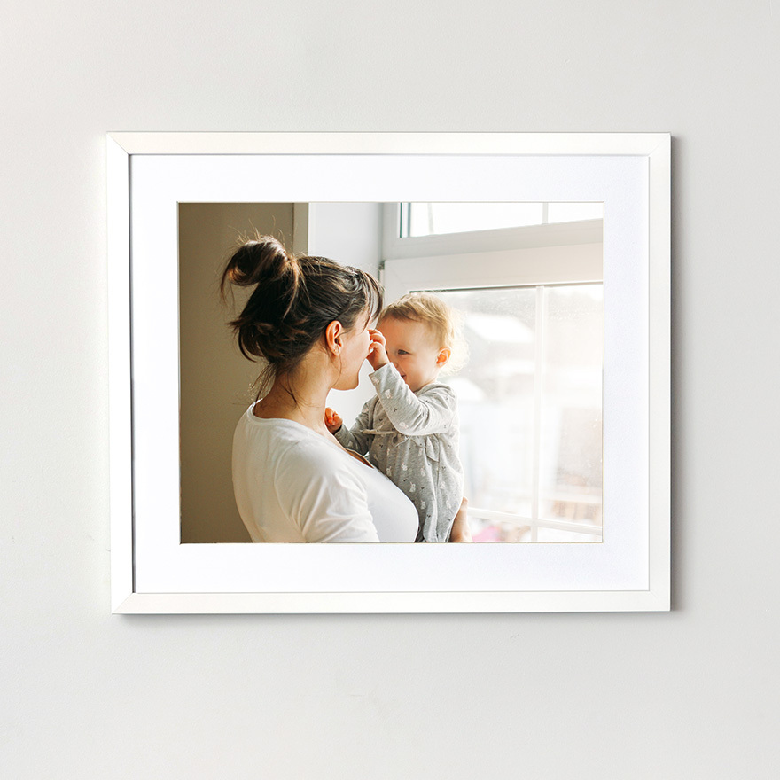 White matted framed photo print showing a parent holding a baby near a window with soft natural light.