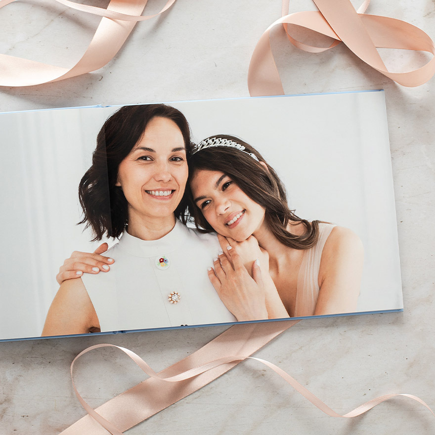 Layflat photo book open to full-bleed portrait of mother and daughter leaning together, styled with ribbon on marble surface.