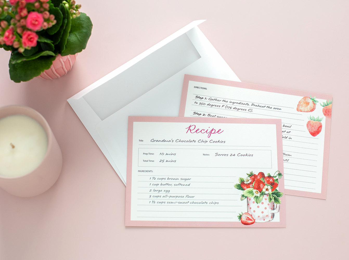 Flat recipe cards with pink border and strawberry illustrations displayed with matching envelope on soft pink background beside candle and plant.