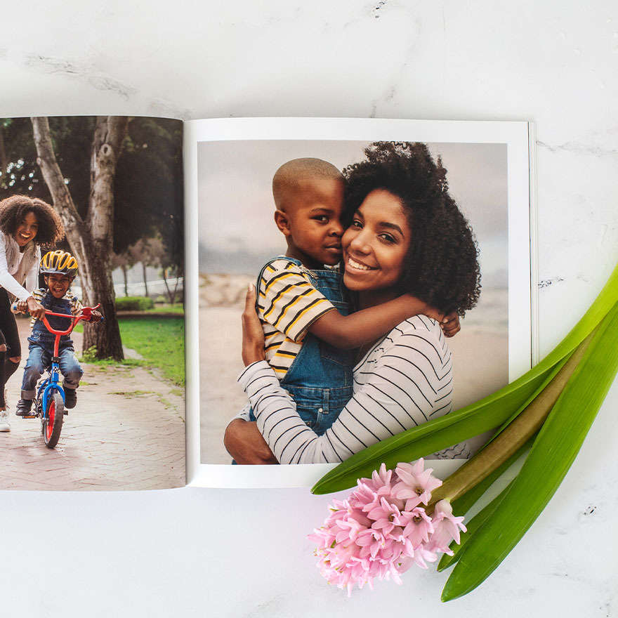 Softcover photo book open to full-page portrait of a mother holding her child, with another family biking photo visible on the opposite page.
