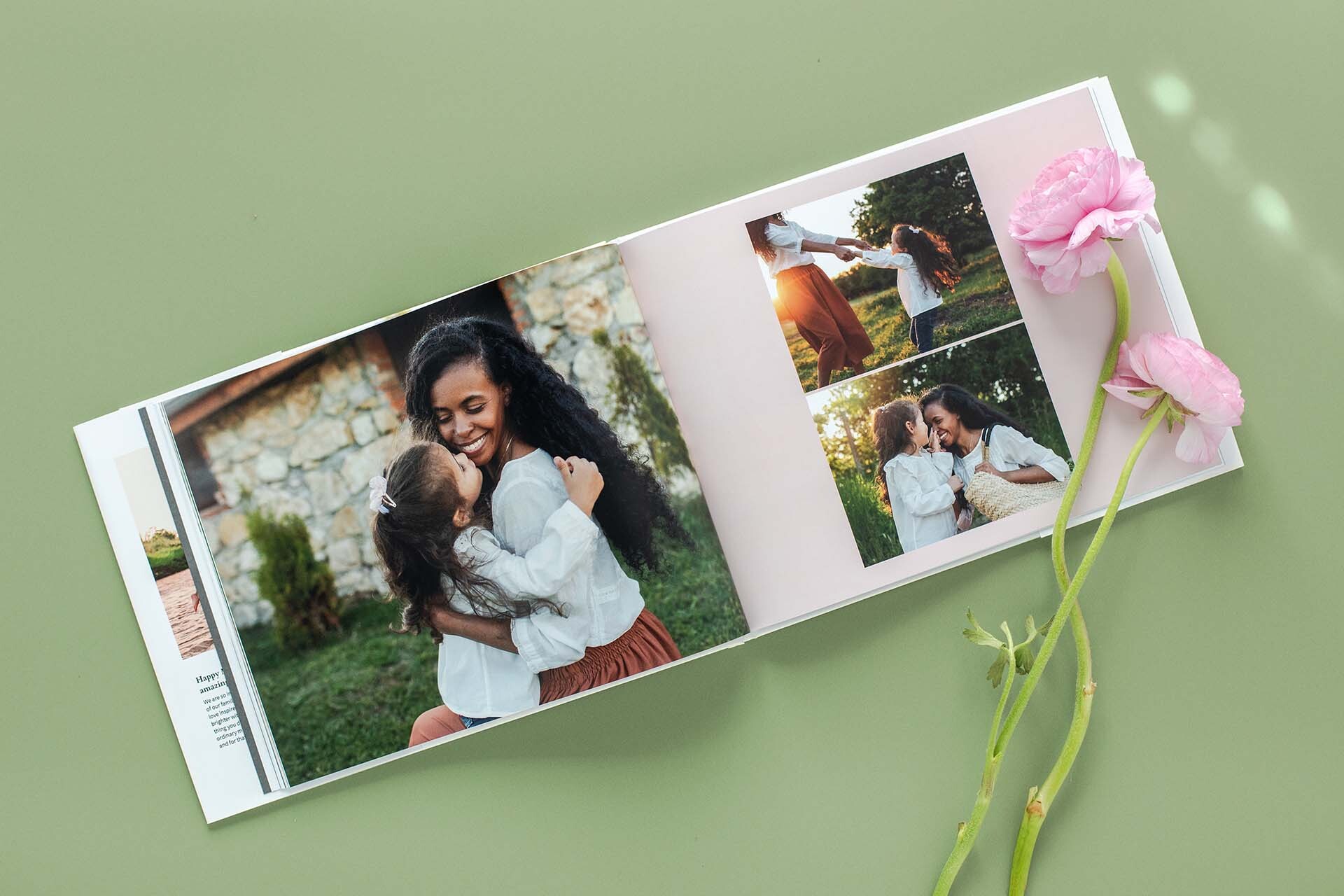 Hardcover photo book open to spread of a mother and daughter hugging and playing outdoors, styled on green background with pink flowers beside the book.
