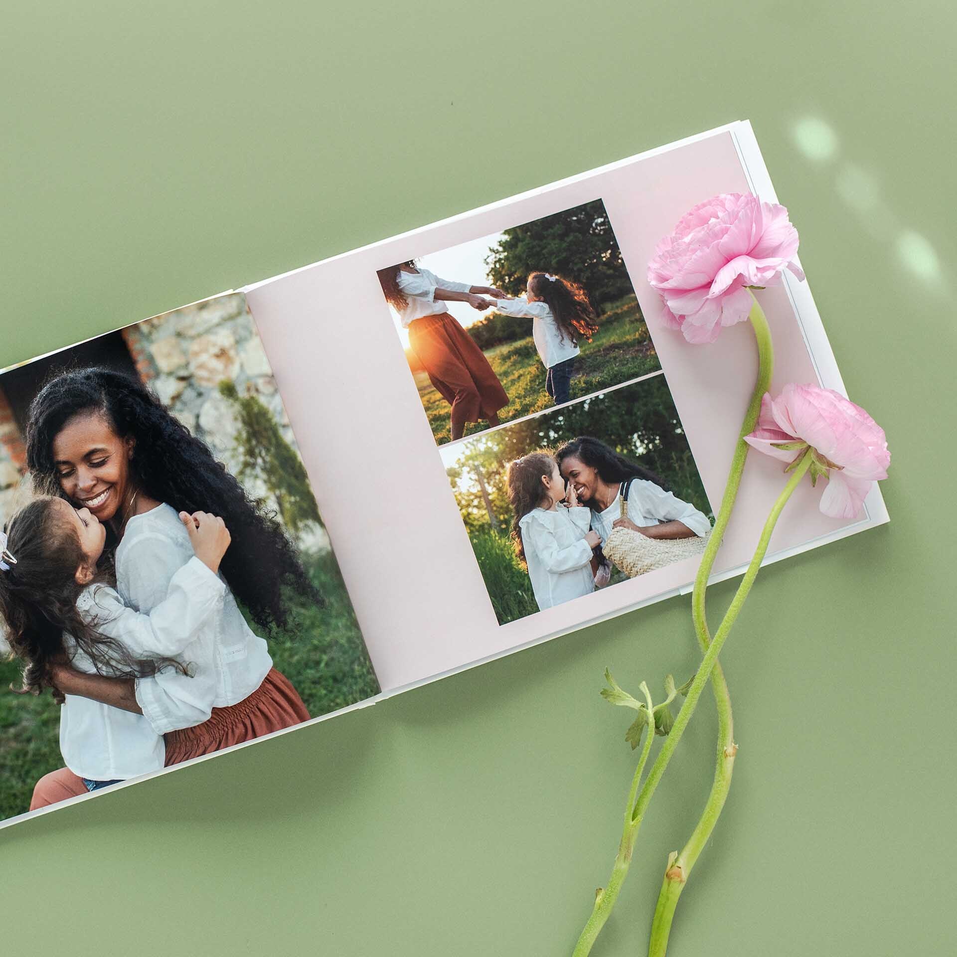Hardcover photo book open to spread of a mother and daughter hugging and playing outdoors, styled on green background with pink flowers beside the book.