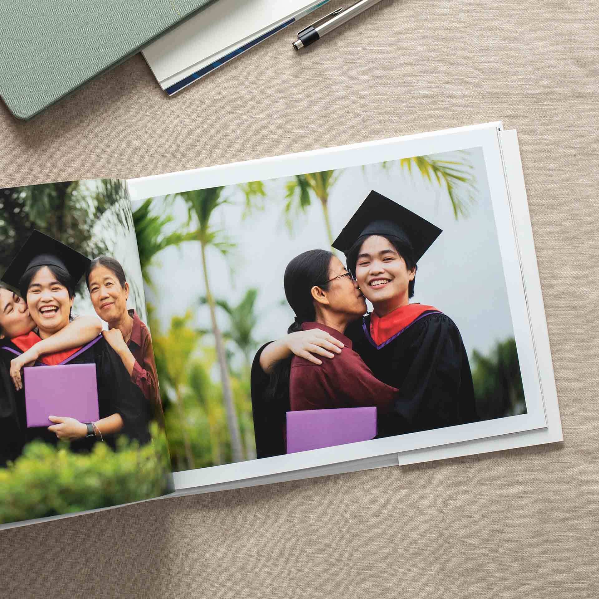 Open hardcover photo book on a table, showing a graduate in cap and gown celebrating with family, including hugs and smiles.