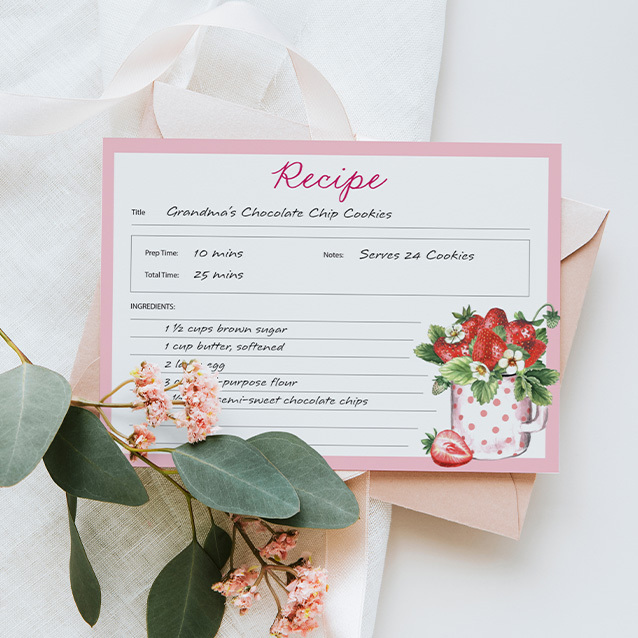 Flat recipe card with pink border and strawberry illustration, printed cookie recipe layout placed on fabric with flowers and leaves.