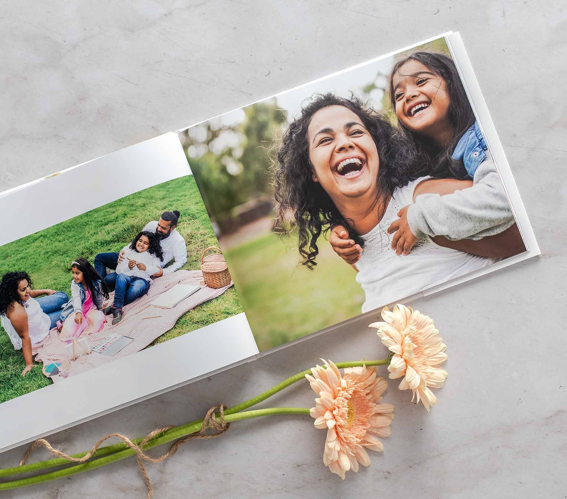 Open hardcover photo book displaying a family picnic scene and a large portrait of a mother and child, styled with peach flowers on a light surface.