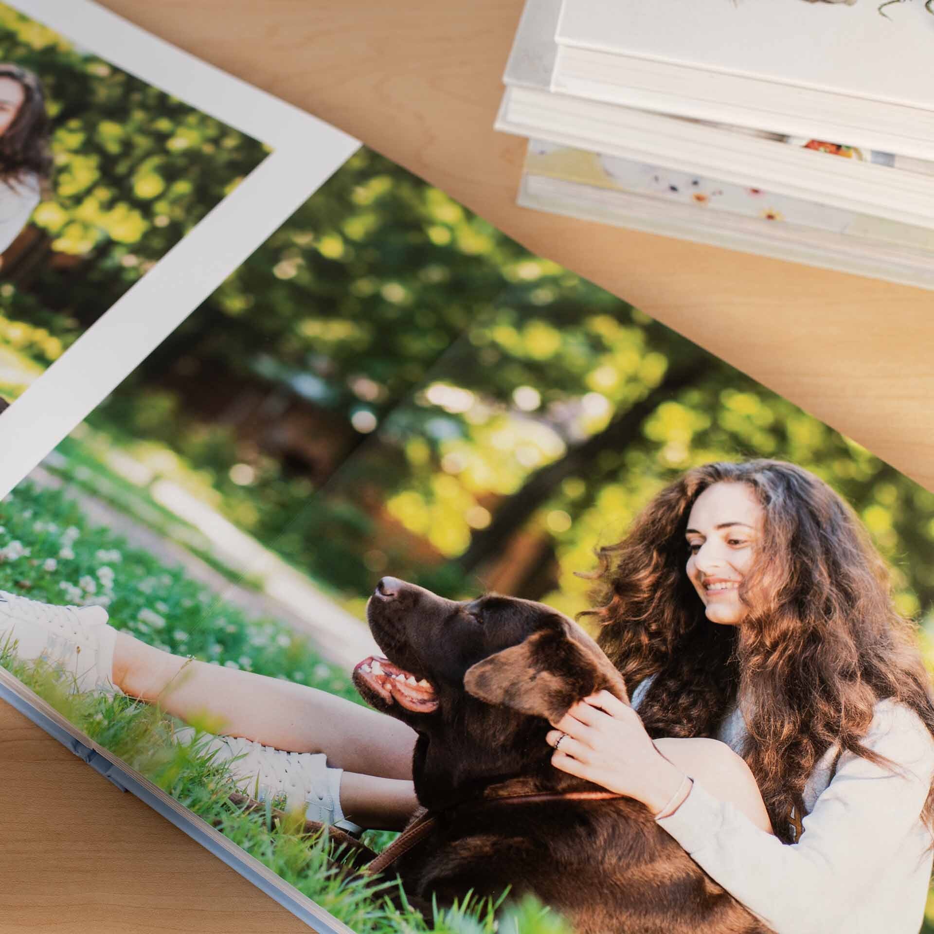 Layflat photo book open on a wooden table displaying a full-bleed image of a person sitting on grass with a dog, next to a coffee cup and stacked books.
