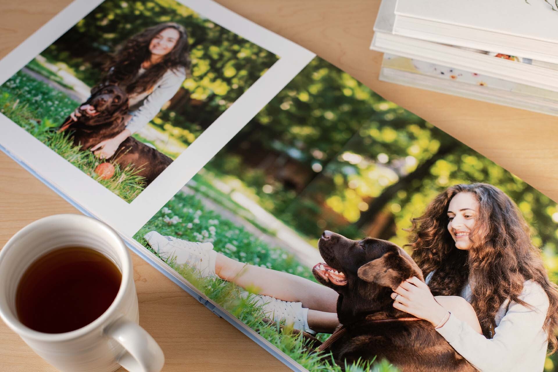Layflat photo book open on a wooden table displaying a full-bleed image of a person sitting on grass with a dog, next to a coffee cup and stacked books.