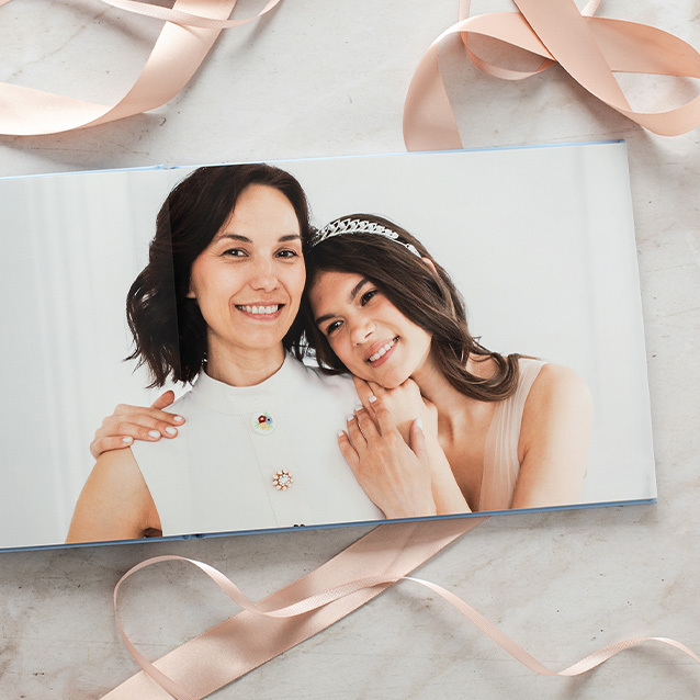 Open layflat photo book featuring a close-up portrait of two women smiling, styled on a marble surface with soft pink ribbon accents.