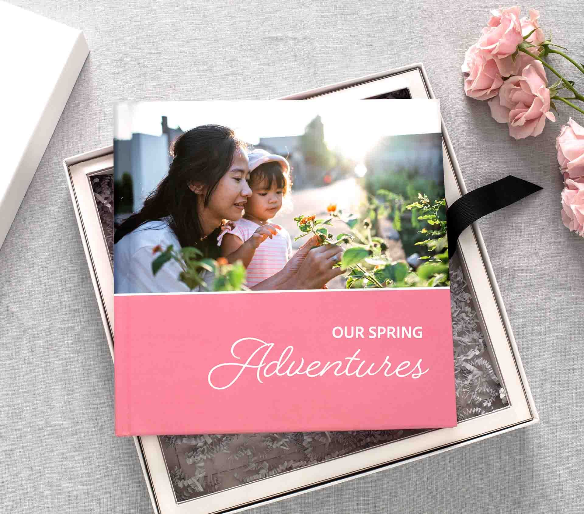 Layflat photo book with pink cover titled “Our Spring Adventures,” displayed in a keepsake box with tissue paper and pink roses.