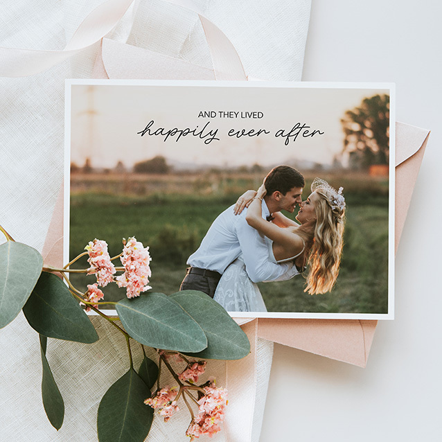 Flat photo card featuring a couple embracing outdoors with printed text reading “And they lived happily ever after,” styled with envelopes and pink flowers.