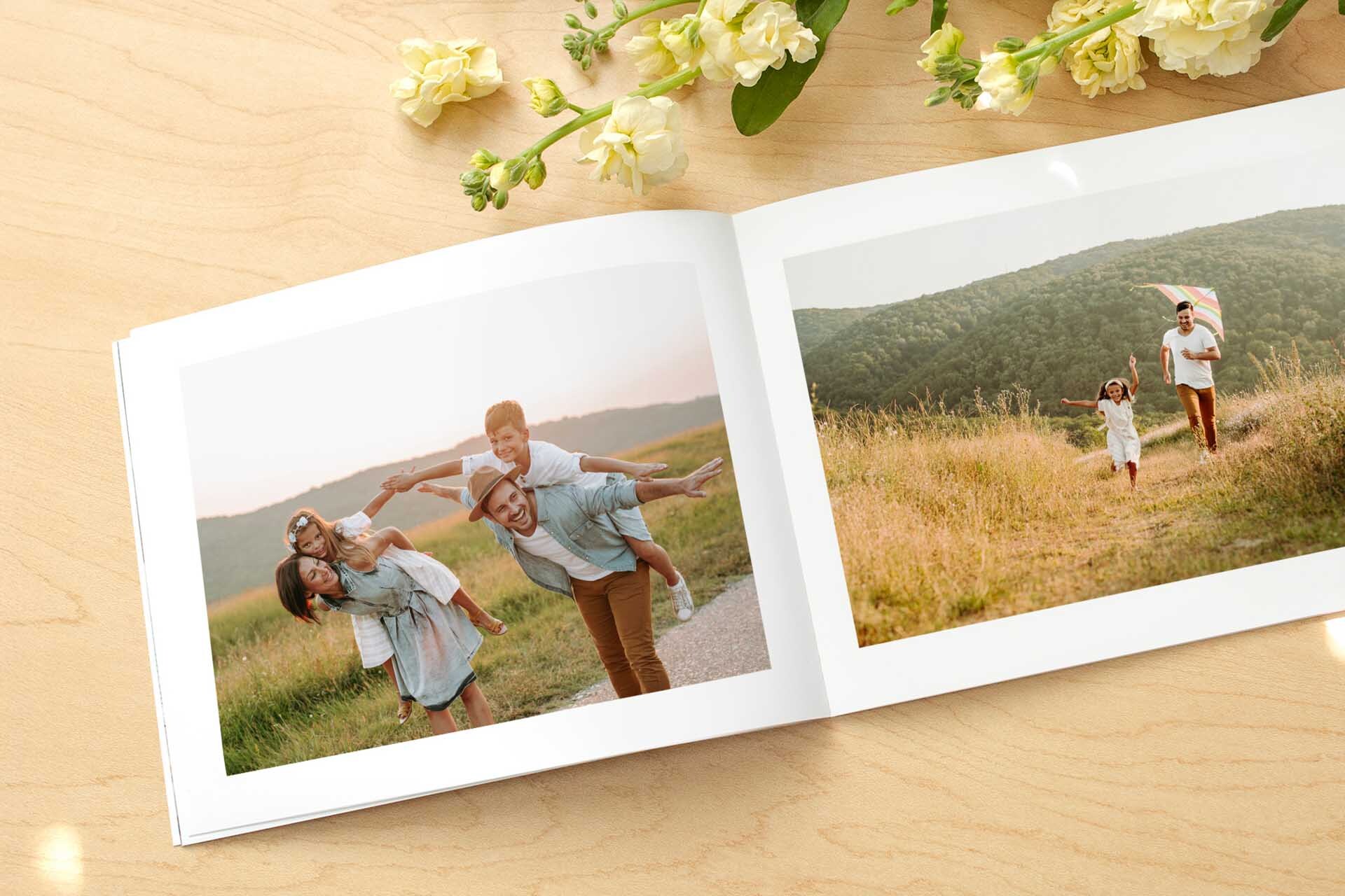Open softcover photo book showing a family playing outdoors at sunset, with flowers on a wooden surface.