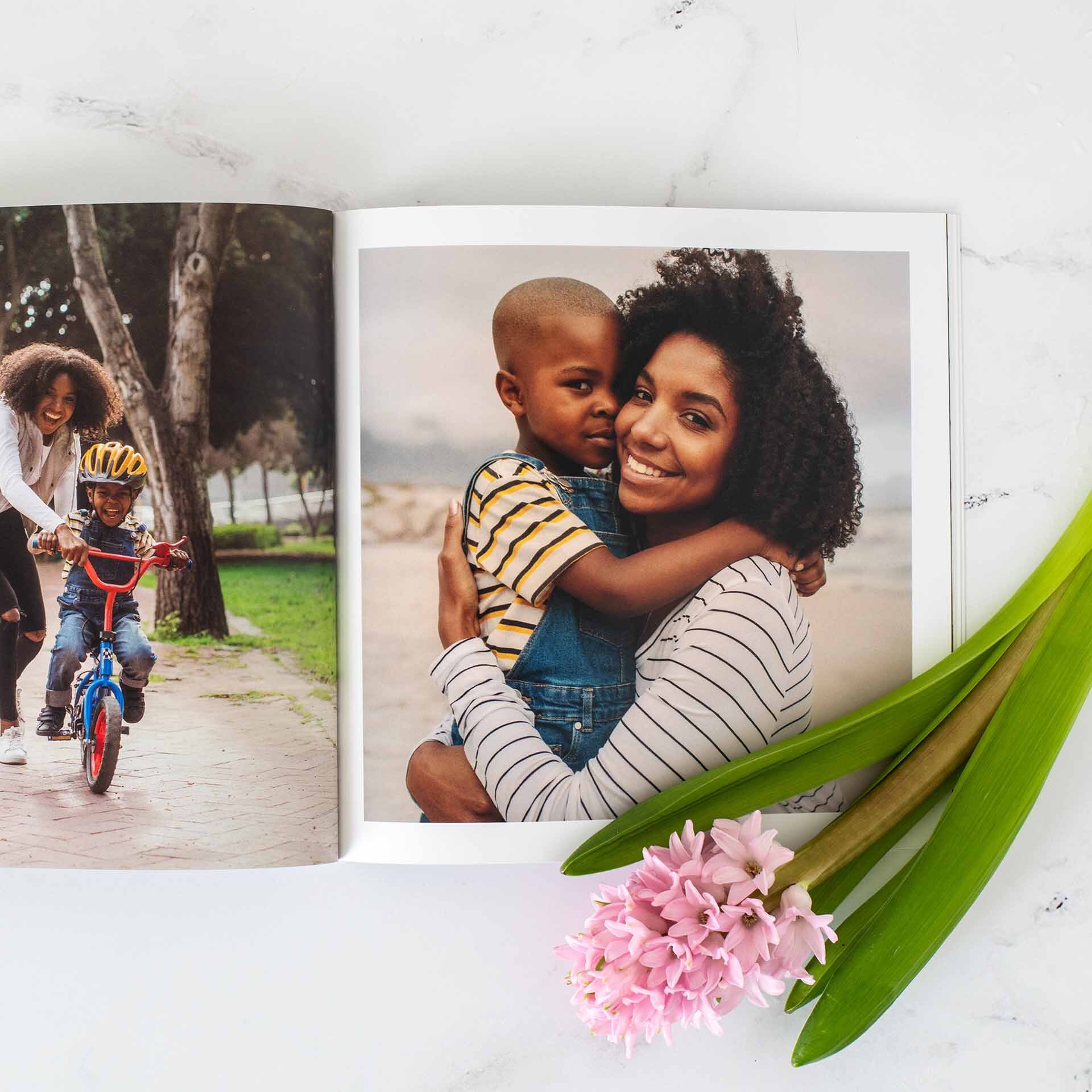 Open photo book showing a mother hugging her young son, with another page of a child learning to ride a bike, styled with pink flowers beside the book.