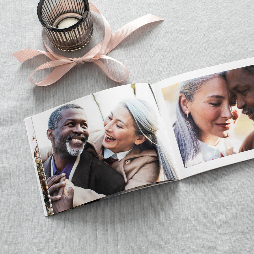 Open softcover photo book on a gray linen surface showing affectionate couple portraits, with a pink ribbon and glass candle nearby