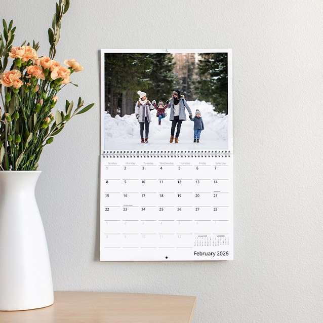Spiral-bound wall calendar open to February 2026, showing a family walking through a snowy forest, hanging beside a vase of flowers