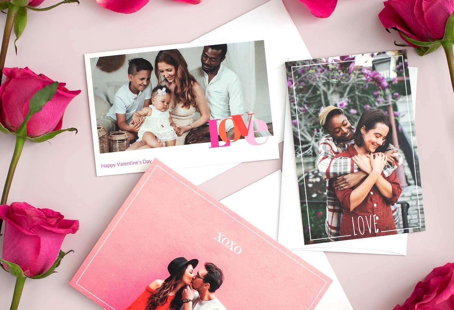 Three custom Valentine's Day photo cards displayed on a soft pink surface with roses—two flat cards featuring romantic and family moments, and one folded card with a couple embracing under blooming trees