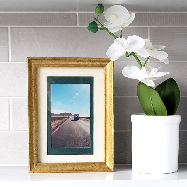 Framed print of a road trip photo in a gold frame next to a white orchid plant on a shelf