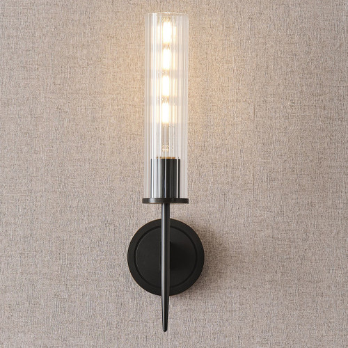 Front view of an illuminated Aicken Sconce Bronze wall light, showing light shining through the clear cylindrical glass shade and the metal base and arm with a brushed bronze finish.