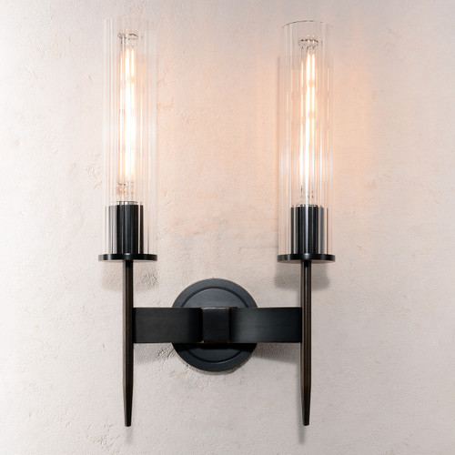 Front view of an illuminated Aicken Double Sconce Bronze double wall light, showing light shining through the clear cylindrical glass shades and the metal base and arms with a brushed bronze finish.