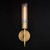 Front view of an illuminate Aicken Sconce Brass, showing the cylindrical 
glass shade and brass circular backplate.