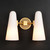 Front view of an illuminated Bianco Double Sconce Brass wall light, showing two glowing milky white conical glass shades and a metal base and arms with a burnished brass finish.