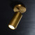 An illuminated Cameron Adjustable Flushmount Brass light, angled downwards showing the cylindrical head with a burnished brass finish and light emitting from it.