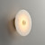 Side angle view of the Daisy Round Alabaster Sconce wall light, showing it mounted on the wall, with the circular alabaster and central metal detail emitting a warm glow when illuminated.