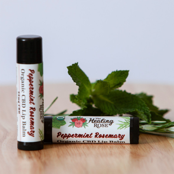 The Healing Rose Peppermint Rosemary Organic CBD Lip Balm tubes beside fresh peppermint and rosemary sprigs