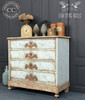 Dixie Belle Paint Putty Chalk Mineral Paint is the perfect paint for any DIY project! A 4 drawer dresser with an old world effect featuring a grey blue color as a base and neutral putty on the bottom and around the sides with wooden appliques underneath each piece of hardware presented with a rust effect. This piece is staged with numerous antique items such as scale, basket with glass water jug painted blue and old seasoning containers.