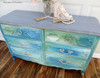 Dixie Belle Paint Voodoo Gel Stain (Water-Based).  A blue, green, and white blended chalk painted dresser with gold handles and a bluish-gray stained top.  White beads with gold tassels are draped over a white figurine and a glass vase are on top of the dresser.  The dresser sits on a brown wood floor, in front of a white wall.