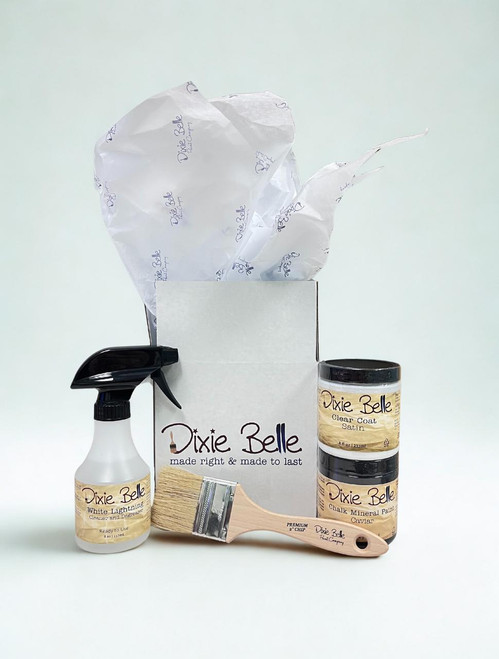 A set of Dixie Belle paint products, including a spray bottle, one small jar of Caviar paint, a paint brush, and a jar of clear coat, displayed in front of an open box with tissue paper.