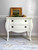 White chalk painted dresser with black handles. Staged with greenery and a gold picture frame on top and book pages strewn on the floor.