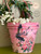 A pink Flower pot. Versailles Garden Transfer is seen applied around the pot. A blurry Wreath in the back ground above the pot with white flowers on the rim of the flower pot.