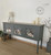 A vintage-style sideboard painted in dark grey with floral designs on its doors. It is positioned against a plain white wall. The sideboard is decorated with a lamp and two candles.