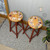 Two wooden stools with round, light brown tops and red legs are adorned with floral wreaths. The stools are placed on a patterned rug in a cozy indoor setting with a plant and a logo in the background.