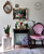 A cozy, vintage-inspired corner featuring a pink tufted chair, a small cabinet adorned with floral and foliage designs, potted plants, a wall mirror, and a floral painting.