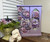 A lavender jewelry armoire adorned with floral designs sits on a wooden surface. To its left is a decorative floral arrangement in a green vase, with moss at its base. In front of the vase, there are white ceramic bunny figurines hugging.
