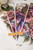 Three transparent sheets, each with a detailed, colorful floral design, are fanned out on a white surface. A wooden stick labeled "Dixie Belle" rests on top. A white flower is partly visible in the top left corner.