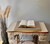 A vintage sewing table with ornate gold accents holds an open book on top of a cane webbing decoupage paper. Tall dried grass decorates the left side, and a fluffy rug covers the floor beneath the table.