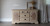 A beige sideboard with decorative trim and four central drawers, flanked by two cabinet doors with a grasscloth decoupage paper. On top are decorative items: a vase with dried grasses, a picture frame, and an orb sculpture.