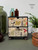 A small chest of drawers with botanical and insect illustrations, including butterflies, bees, and plants. It sits on a table surrounded by glass bottles, a green vase, a potted fern, and a wooden object.