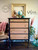 A vintage dresser with light wood drawers and dark frame is topped with books, globes, and a clock. An empty ornate frame hangs above. A plant, pink scarf, and hat decorate the scene. Logos on the wall read “Dixie Belle Paint Company Content Creator” and “by Melinda MTZ Crew Designs”.