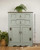 A vintage mint-green cabinet with two drawers and two doors, featuring ornate metal handles and decorative carvings, stands on a wooden floor with green plants on and beside it. A watermark reads “GRACIE’S HOUSE Painted Furniture.”.