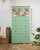 A mint green dresser with four drawers, the top drawer decorated with a floral decoupage paper featuring large pink flowers. The dresser stands on a wooden floor, surrounded by plants and a wicker basket.