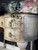 Dixie Belle Paint Van Dyke Brown Glaze. The right side of a light brown chalk painted des with darker brown edges and gold handles.  A green plant in a pot sits on top of the desk and a floral picture is in the background.