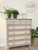 Dixie Belle Paint Putty Chalk Mineral Paint is the perfect paint for any DIY project! A muted taupe colored dresser featuring 2 short drawers next to each other on top with a basket weave stencil and 4 long skinny drawers underneath with a rattan like pattern surrounded by various potted greenery.