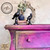 A vintage dresser with a gradient of purple and gold hues is adorned with two black bird figurines, a small vase with leaves, and stacked books. An ornate mirror is placed behind them. The image showcases the mix of Gold and Bronze Gilding wax.
