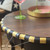 A round wooden table with a scalloped edge painted in alternating gold and black stripes. Two glasses with green stems and a pink tray sit on top. An arrow points to the edge labeled "Gold Gilding Wax."