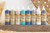 Dixie Belle Paint Voodoo Gel Stain (Water-Based).  8 bottles of VooDoo Gel Stain, 1 of each color, sit side by side on top of a piece of brown wood.  White beads and feathers are on the table in front of the wood, a light brown feather is to the left of the stain, and a brown woven basket is behind the stain, to the right.
