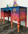 Belles and Whistles Western Boho - Silkscreen Stencil provides an extra decorative touch to any project!  Blue, pink, orange and yellow blended and layered chalk painted desk with the Western Boho Silkscreen Stencil applied to the front and sides of the desk in gold paint.  Drawer handles are gold.  The desk stands on a brown wood floor, in front of a white wall.  On top of the desk are blue, green and purple glass vases, a gold figurine, an orange flower, and a brown pot with greenery inside.