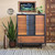A mid-century modern wood dresser with black accents stands against a beige wall, surrounded by green plants in woven pots. Above the dresser hangs a framed painting of cacti under a dark sky. A patterned rug covers the floor.